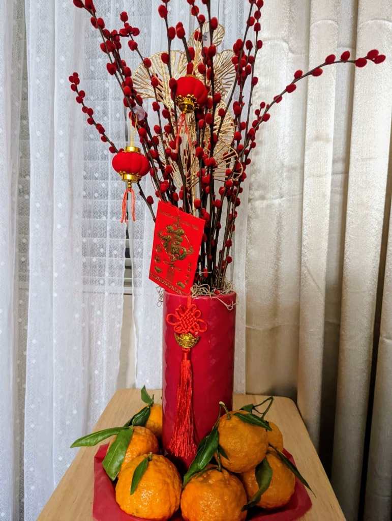Vase of branches with red fuzzy tips, decorated with lunar new year items, and surrounded by oranges